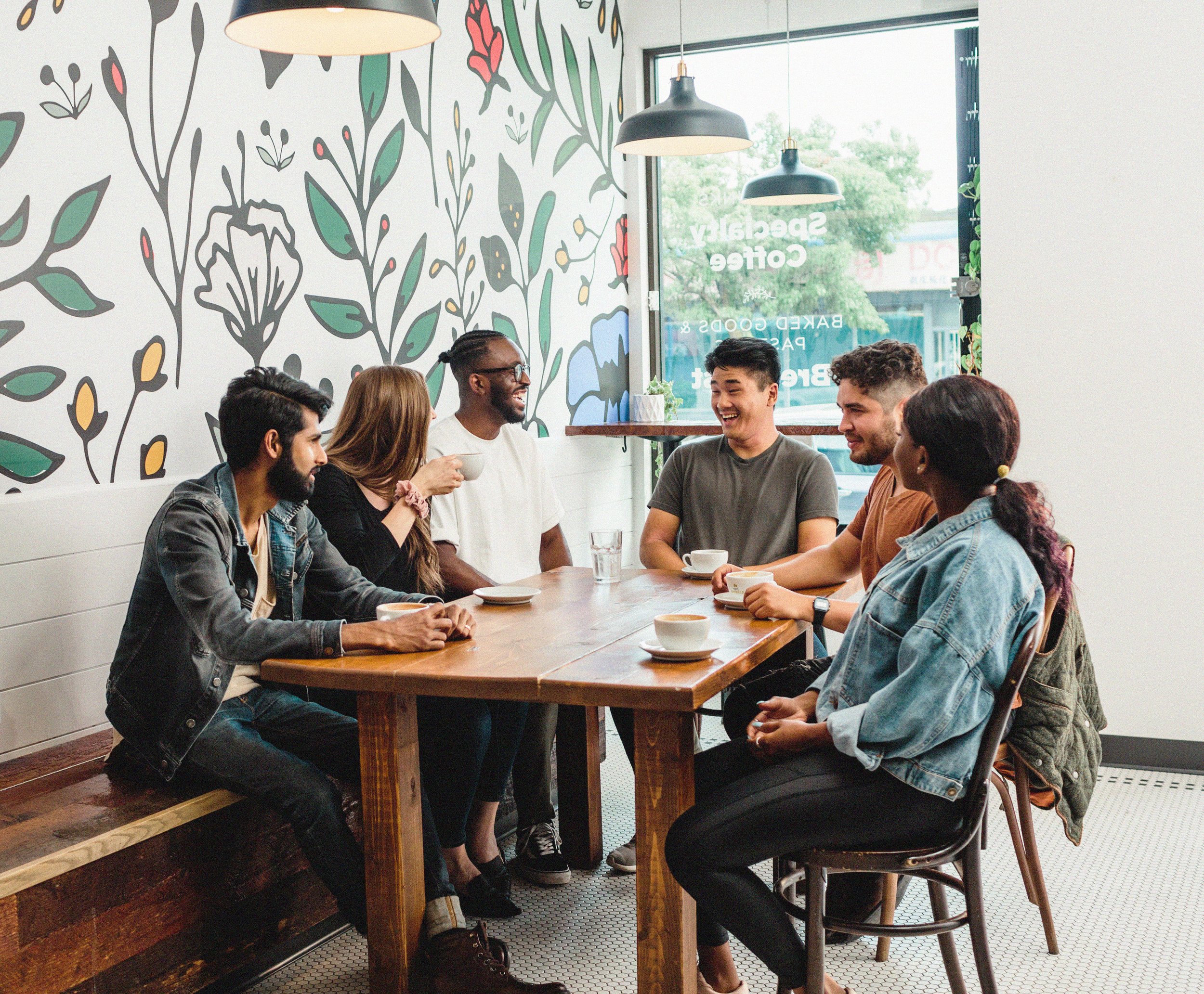 Group of six diverse young adults sitting around a wooden table in a coffee shop, laughing and chatting.