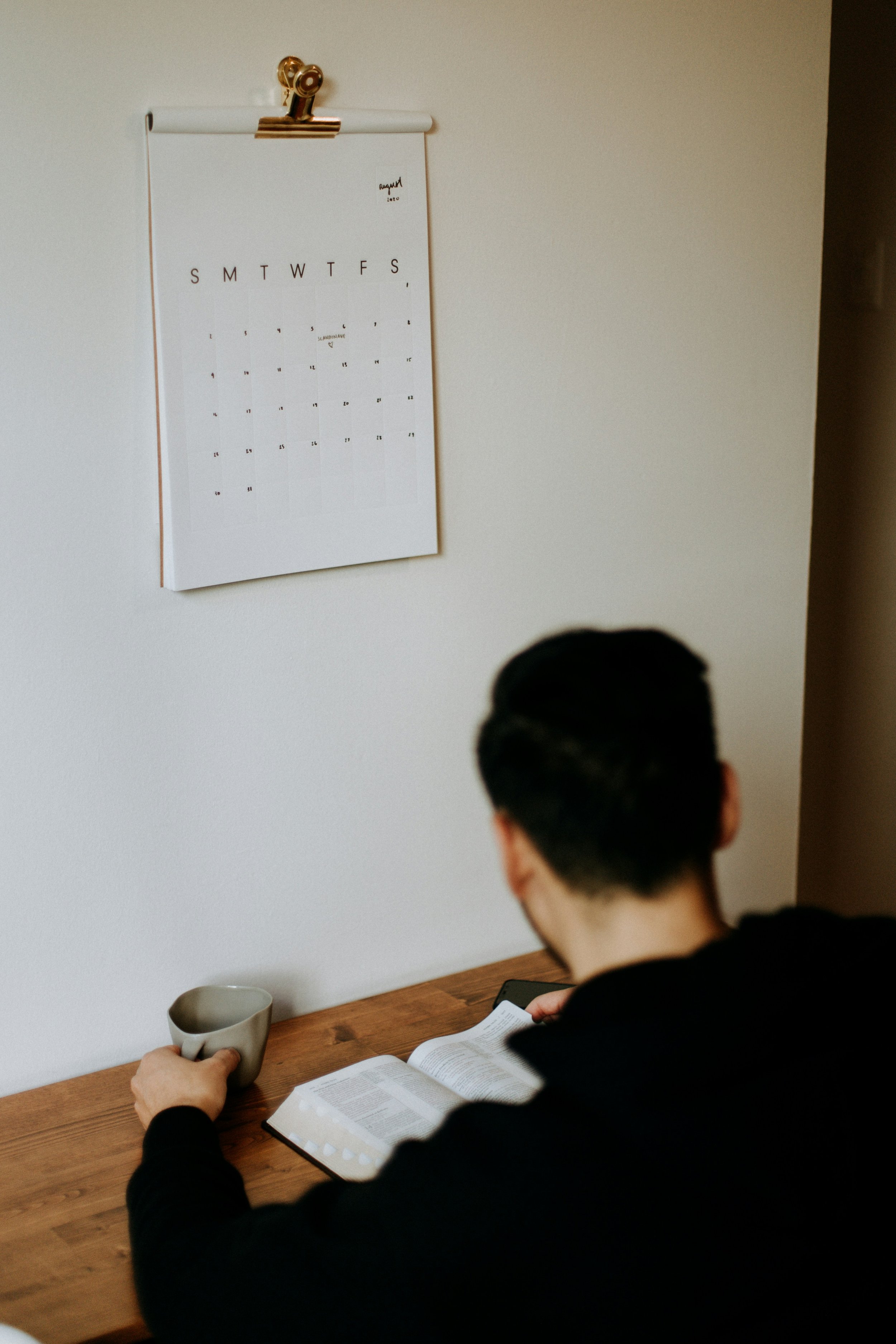 A person sitting at a wooden desk, reading a book, with a cup in hand, and a calendar on the wall.