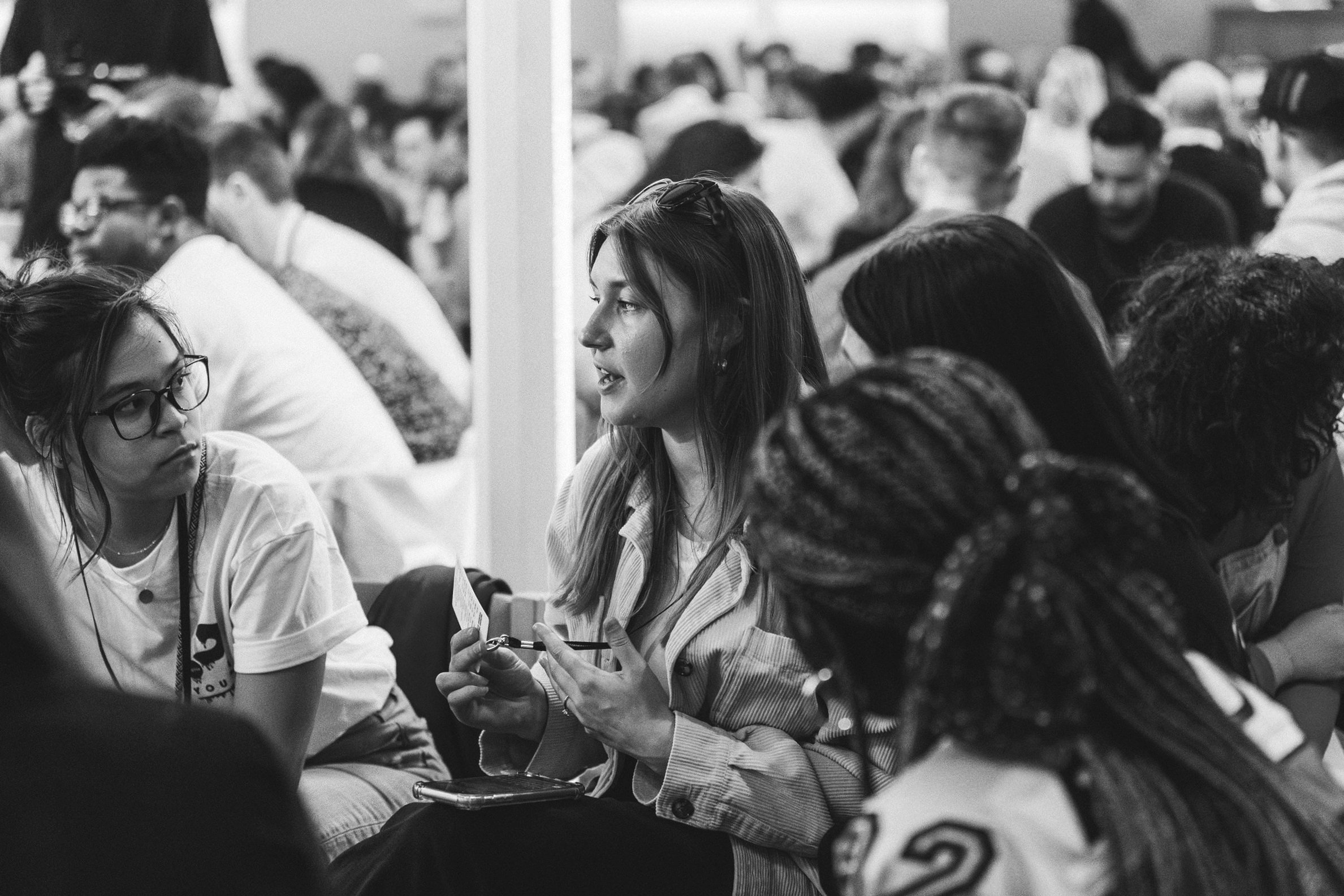 A black and white photograph of a diverse group of people sitting closely together at an indoor event, with one woman in the center holding a pen and speaking.