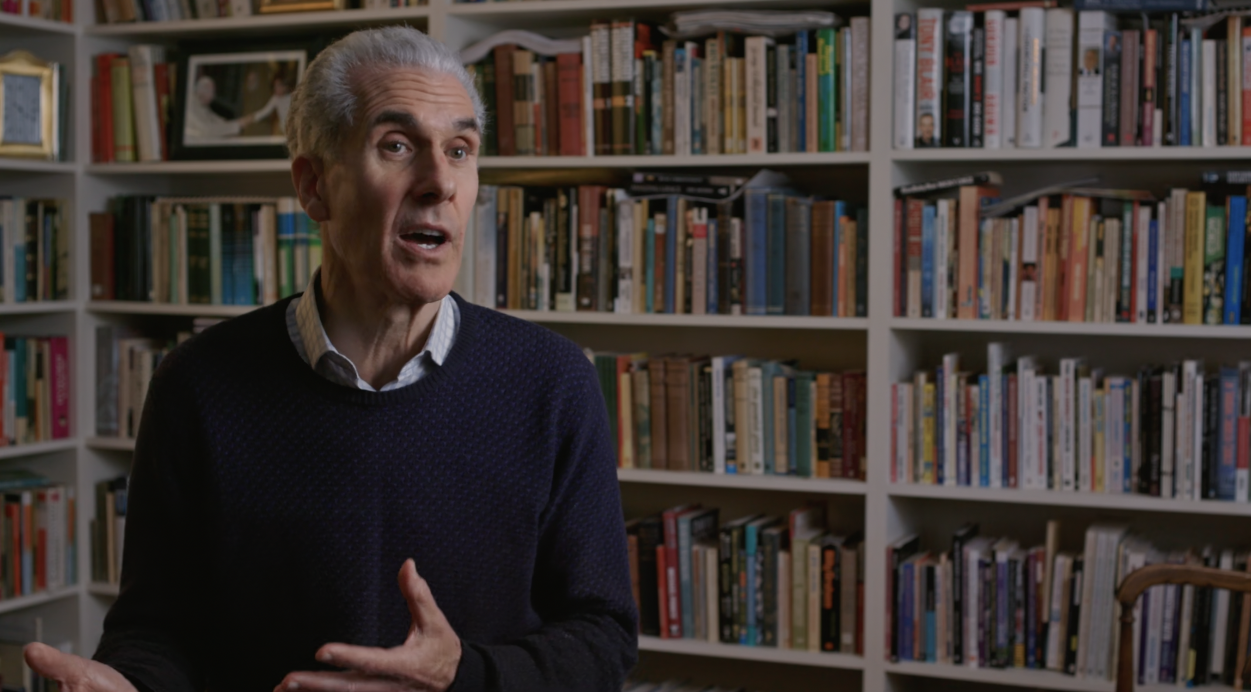 Nicky Gumbel wearing a dark sweater and collared shirt, speaking in front of a bookshelf filled with books.